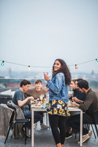 Portrait of smiling woman with drink standing by table during social gathering on terrace building