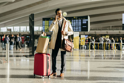 Man with dark hair in stylish clothes talking on phone while standing with suitcase in terminal of airport