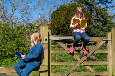 Two girls enjoying reading from yellow and blue, textbook while sitting on wooden fence.