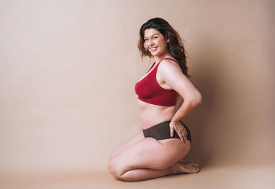 Self loving young woman plus size in underwear sitting on beige background, body love