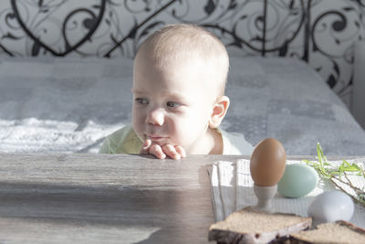 A sad little child sits at a table with bread and easter eggs with light from the window.