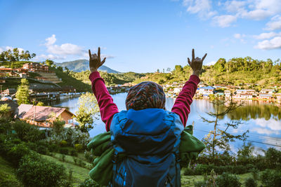 Rear view of woman with arms raised standing against lake by village