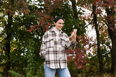 A cute girl in warm clothes has a video call on the phone against the background of an autumn forest