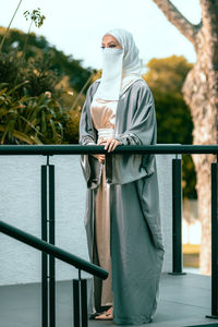 Full length picture of a veiled woman with white niqab and long dress standing against railing