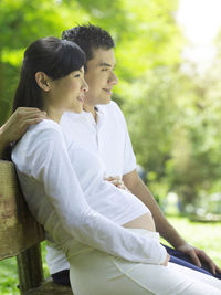 Pregnant woman with husband sitting on bench against tree