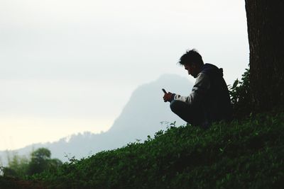 Man using smart phone while sitting on field against sky