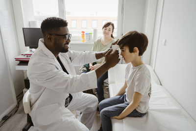 Male pediatrician examining boy sitting on bed in examination room at healthcare center
