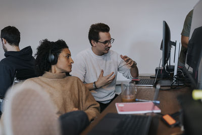 Young male programmer explaining strategy to female colleague by computer at desk in office