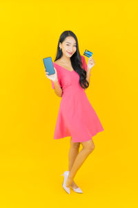 Full length of young woman using mobile phone against yellow background