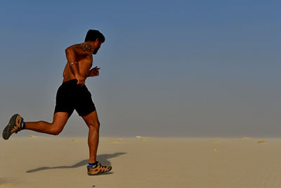 Full length of shirtless young man running on sand at desert during sunny day