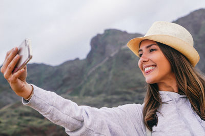 Happy young woman sticking out tongue while taking selfie against mountains