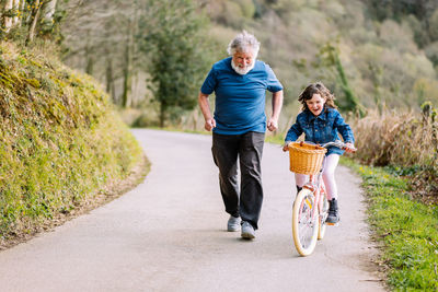 Full body of active grandfather running with cute positive granddaughter riding bicycle on path in countryside with green trees