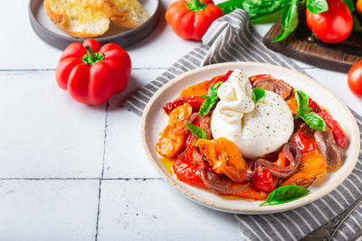 Burrata cheese with baked tomatoes, pepper, red onion and fresh basil on white tile background.