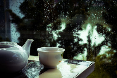 Close-up of teapot with cup on table against wet window