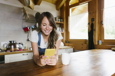 Smiling young woman using smart phone in kitchen at home