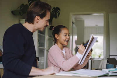 Smiling daughter doing homework while sitting with father at home