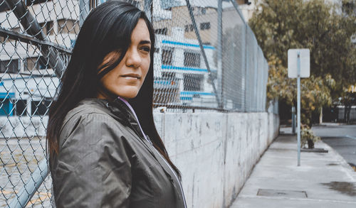 Thoughtful young woman standing on footpath by chainlink fence