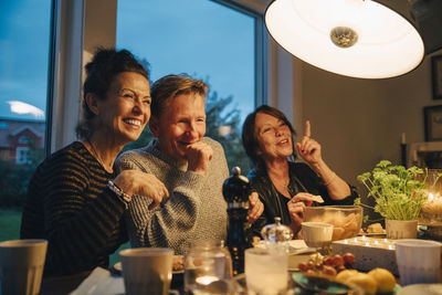 Happy senior women and man sitting at illuminated dining table while enjoying dinner party