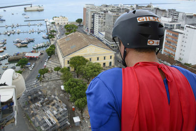 Man wearing black helmet and wearing a hero costume looking down from above the building.