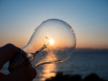 Close-up of hand holding light bulb against sky