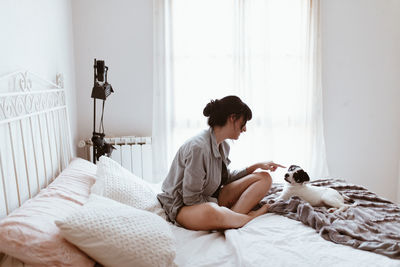 Side view of smiling young woman playing with dog while sitting on bed at home