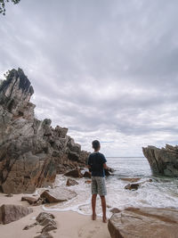 Rear view of man standing on rock at beach against sky