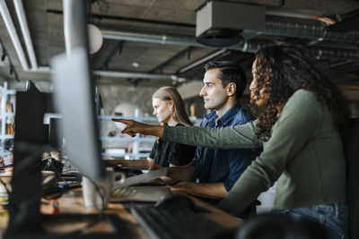 Female coder pointing at device screen while sitting with colleagues in office