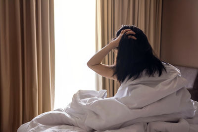 Rear view of woman with hand in hair sitting on bed at home