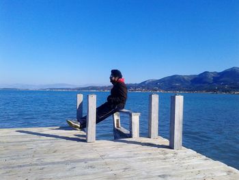 Side view of man sitting on pier over sea against clear blue sky