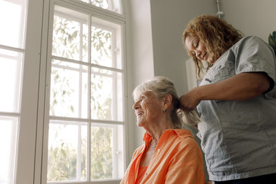 Young female caregiver braiding smiling senior woman's hair at nursing home