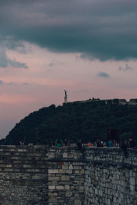 People on wall by buildings against sky at sunset