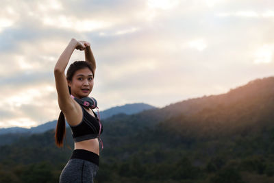 Young woman with arms raised standing against sky during sunset