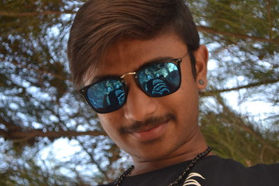 Low angle portrait of smiling young man wearing sunglasses against tree