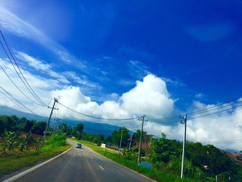 Electricity pylons on road against cloudy sky