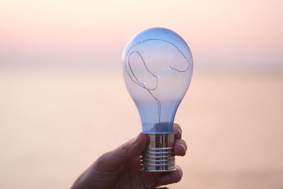 Close-up of hand holding light bulb against sky