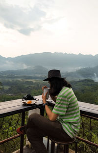 Side view of woman using mobile phone while sitting on mountain against sky