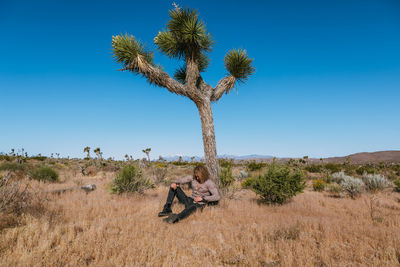 Full length of young man sitting by tree at desert against sky