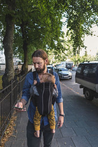 Man using mobile phone while carrying baby in carrier on sidewalk