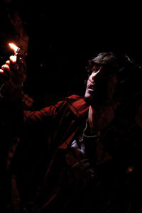 Young man holding lit cigarette lighter against fence at night