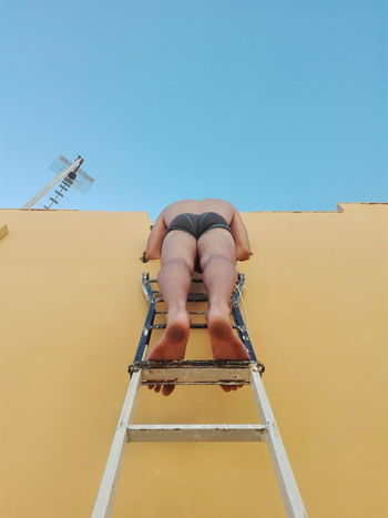 Low section of shirtless man on ladder against clear sky