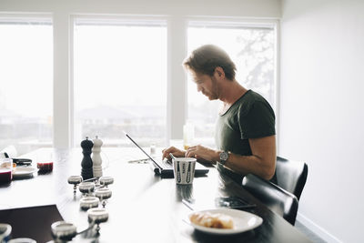 Side view of mature man sitting at dining table using laptop against window