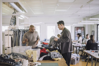 Male fashion designers discussing with each other while sorting recycled clothes at workshop