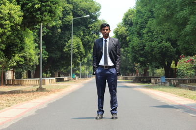 Full length portrait of young man wearing suit standing on road against trees