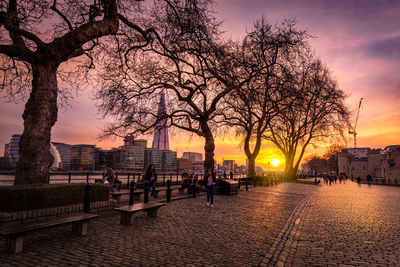 Bare trees by buildings against sky during sunset