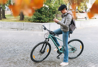 Portrait of young woman in cap and sunglasses using mobile on bicycle on sunny autumn day in city