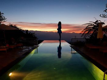 Silhouette woman standing by swimming pool against sky during sunset