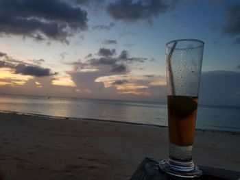 Glass of beer on beach against sky during sunset