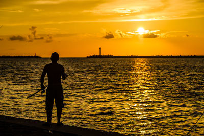 Silhouette man fishing at sea shore against sky during sunset