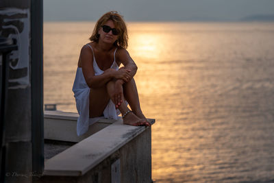 Young woman sitting by sea against sky during sunset