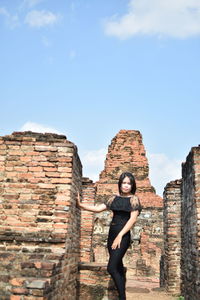 Portrait of young woman standing by old ruin temple against blue sky during sunny day